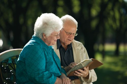 An elderly couple sits on a bench in the park and reads the scriptures together.