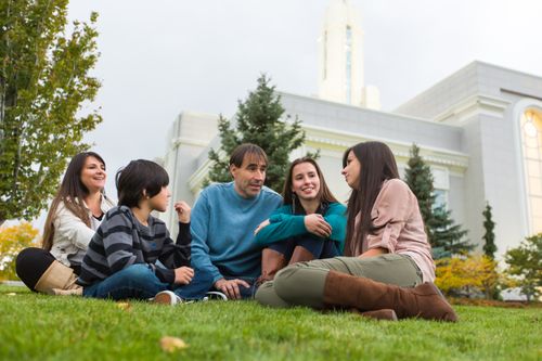 A father, mother, two daughters, and a son sit on the grass near trees and talk outside the Mount Timpanogos Utah Temple.