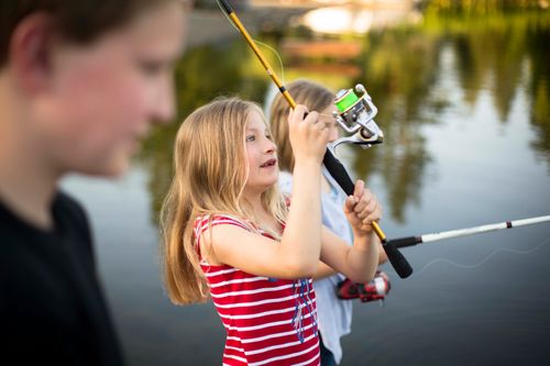 A young girl with light brown hair and a red and white striped shirt holding a fishing pole up in the air by a lake.