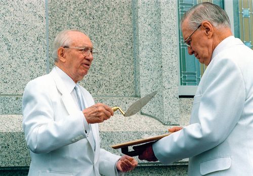 Gordon B. Hinckley and another man stand in all white against a granite temple wall. Hinckley holds a trowel in his hand while the other man holds out some other tool toward President Hinckley.