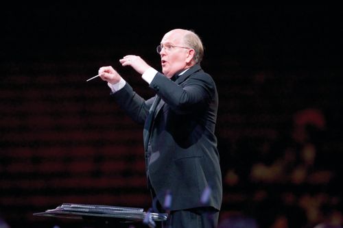 Mack Wilberg in a black suit, standing behind a music stand and conducting the choir with both arms.