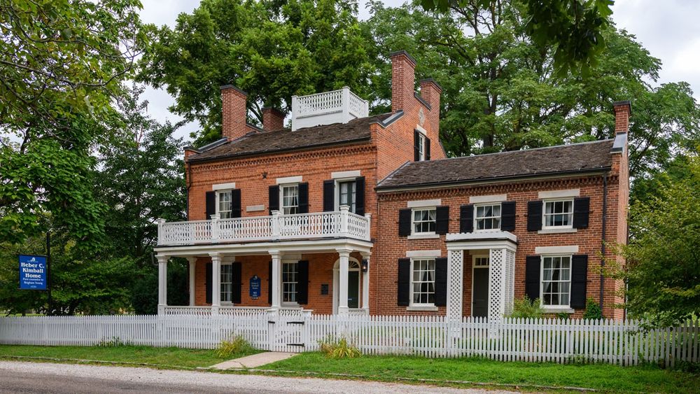 A two-and-a-half story brick home with a double white porch and sizable addition to the right with a white picket fence.