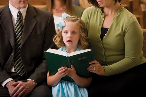 A young girl with a bow in her hair and a light blue dress sits between her parents while singing from a hymnbook.