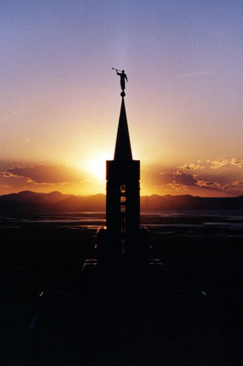 A view of the Bountiful Utah Temple spire, with the angel Moroni silhouetted against an orange sunset in the background.