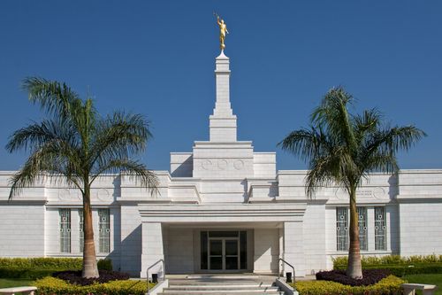 The front entrance to the Oaxaca Mexico Temple, with a clear blue sky overhead and two large palm trees near the doors.