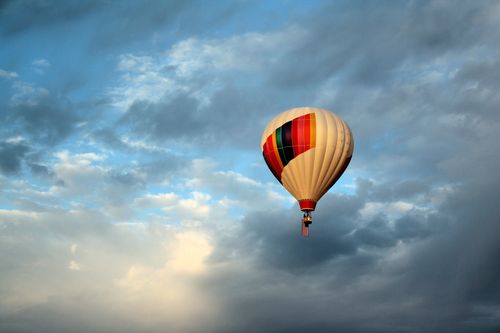 A white hot air balloon, decorated in colored stripes, is seen flying through white and gray clouds, with blue sky poking through the clouds.