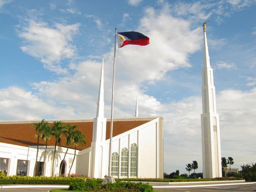 A view of the entrance and grounds to the Manila Philippines Temple, with a separated spire tower to the right and the Philippines flag blowing in the wind on a sunny day.