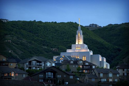 The Draper Utah Temple at night from afar, with some of the neighborhood houses seen in the front of the image and a green mountain in the background.