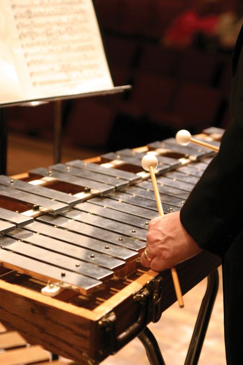 A man holding mallets while playing a glockenspiel and looking at sheet music on a metal music stand.