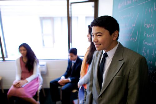 A young man in a suit and tie stands at the front of a classroom next to a chalkboard and talks to a room of young men and young women.