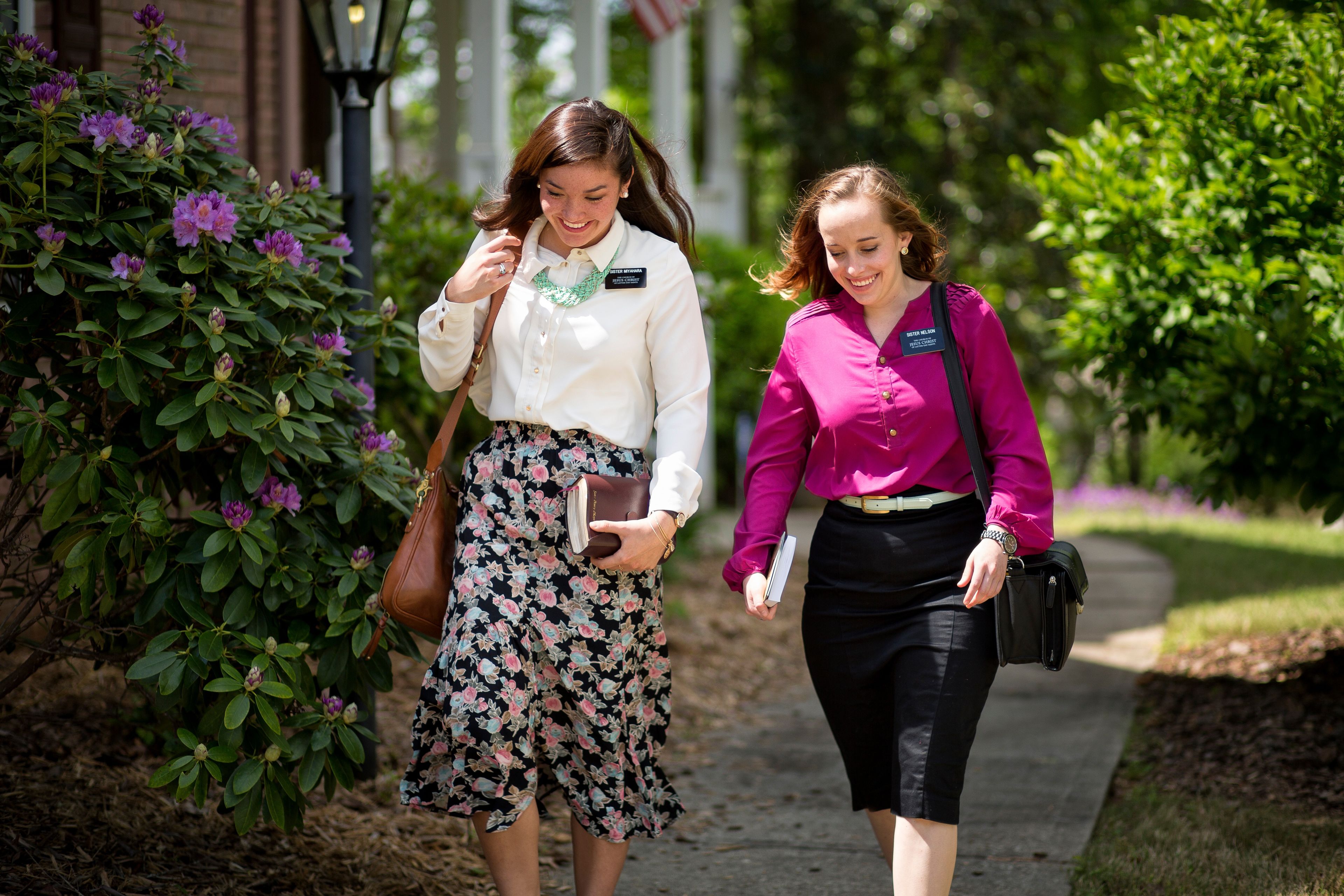 Two sister missionaries carrying bags and scriptures while walking down a path.