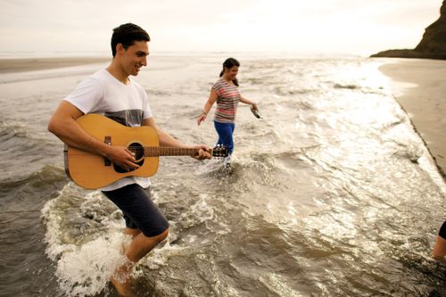 A young man walks in the water along the shore with a young woman while he plays his guitar.