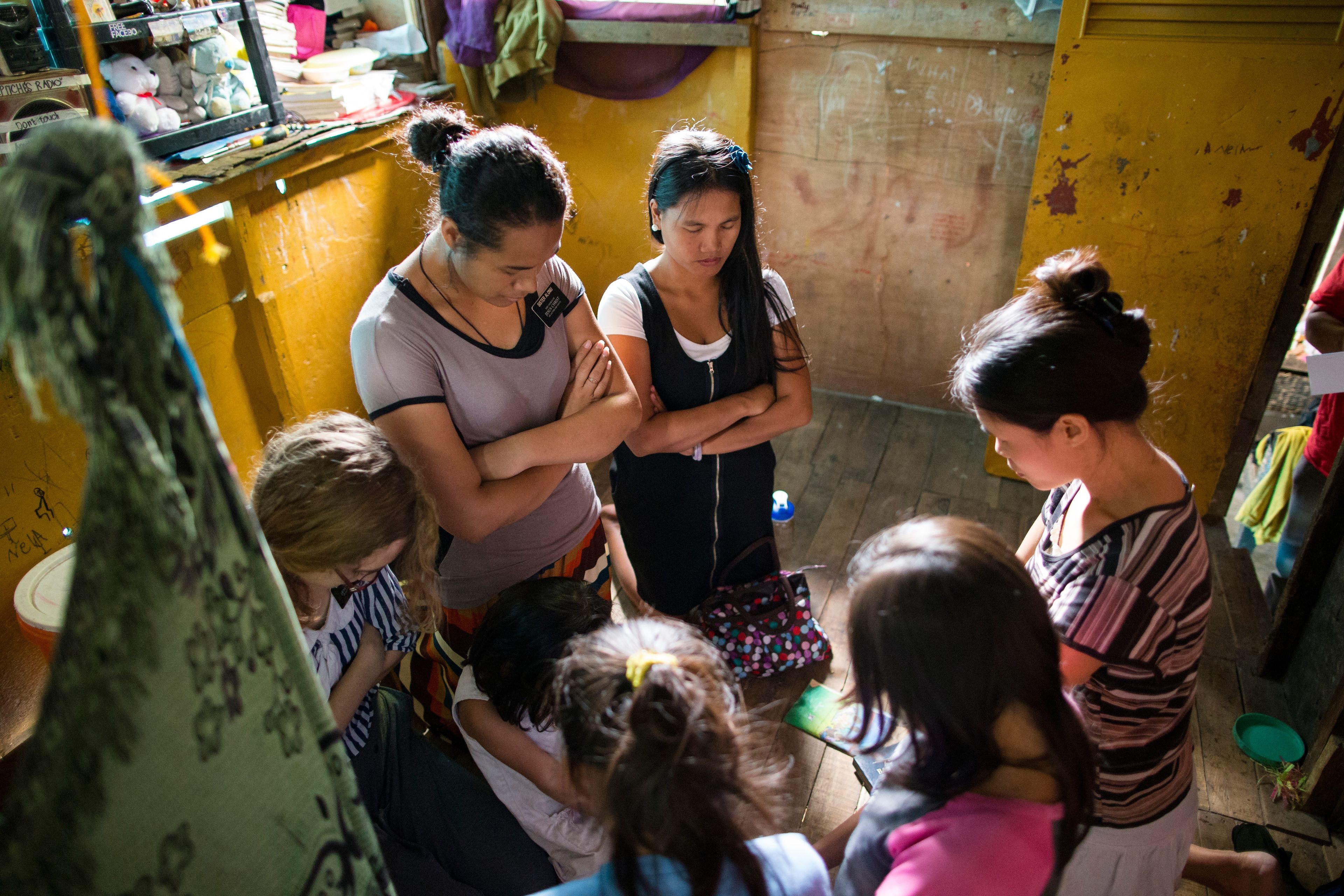 Three sister missionaries kneeling in prayer with a family in the Philippines.