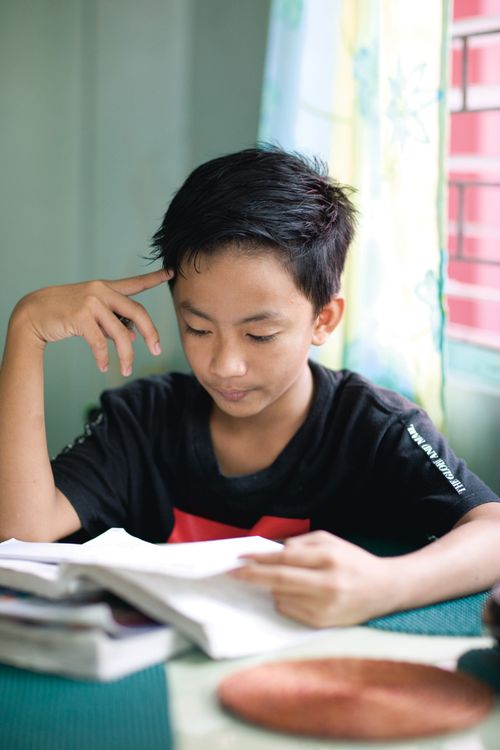 A young boy with short black hair sits at a table in his home and reads from a textbook, with a pile of books on the table.