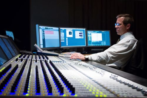 A man in a white shirt sits adjusting a soundboard next to three monitor screens.