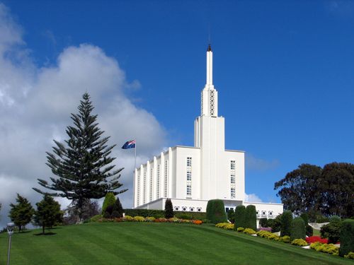 The Hamilton New Zealand Temple, with a neatly manicured green lawn and a thin white cloud in the blue sky to the left of the temple’s spire.