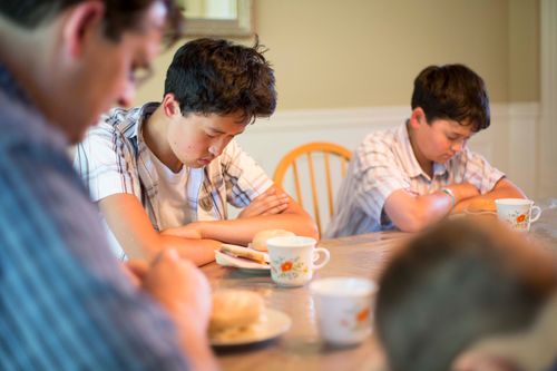 A father leads his family in prayer at the dinner table while his three sons bow their heads and fold their arms.