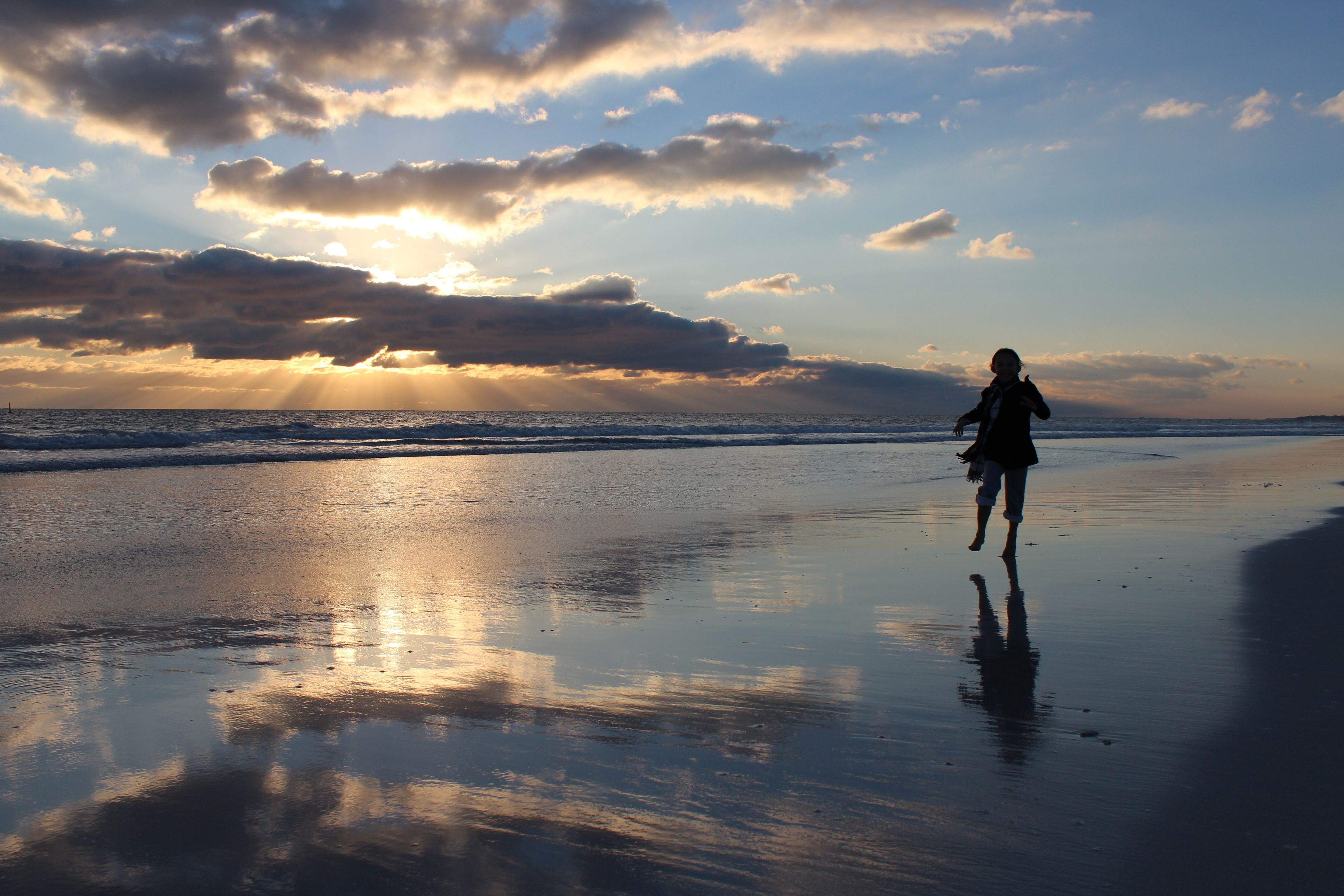A silhouette of a woman walking on the shore with the sun setting behind her and reflecting on the water.