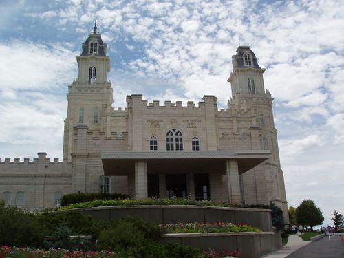 The front entrance of the Manti Utah Temple, with a view of the grounds and flowers and with clouds spotting a blue sky in the distance.