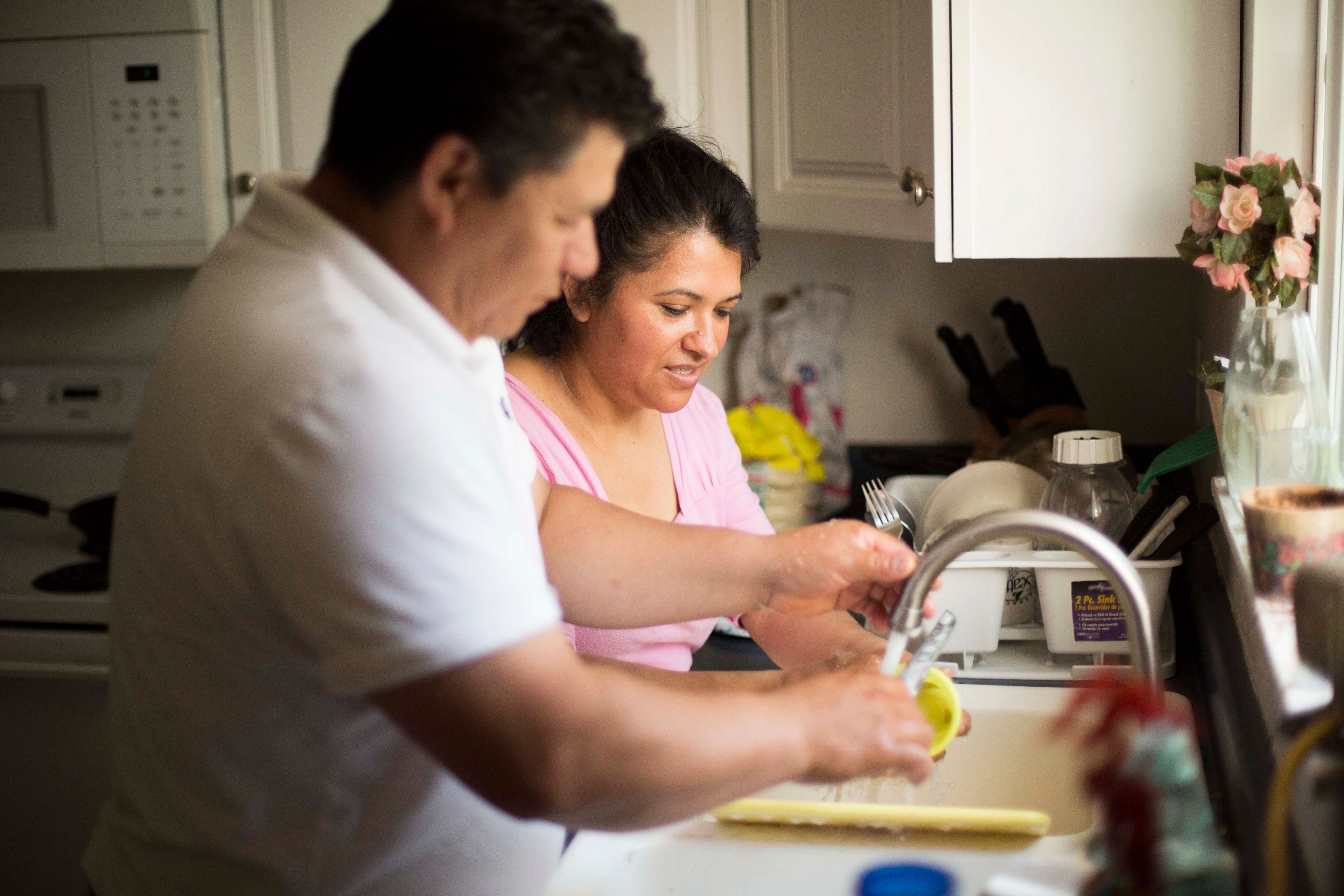 A husband and wife stand by their kitchen sink and wash dishes together.