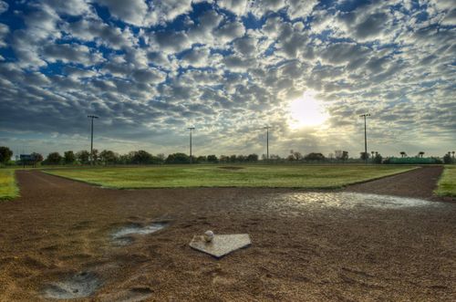 A baseball field with a baseball lying on home base and the sun shining through the clouds.