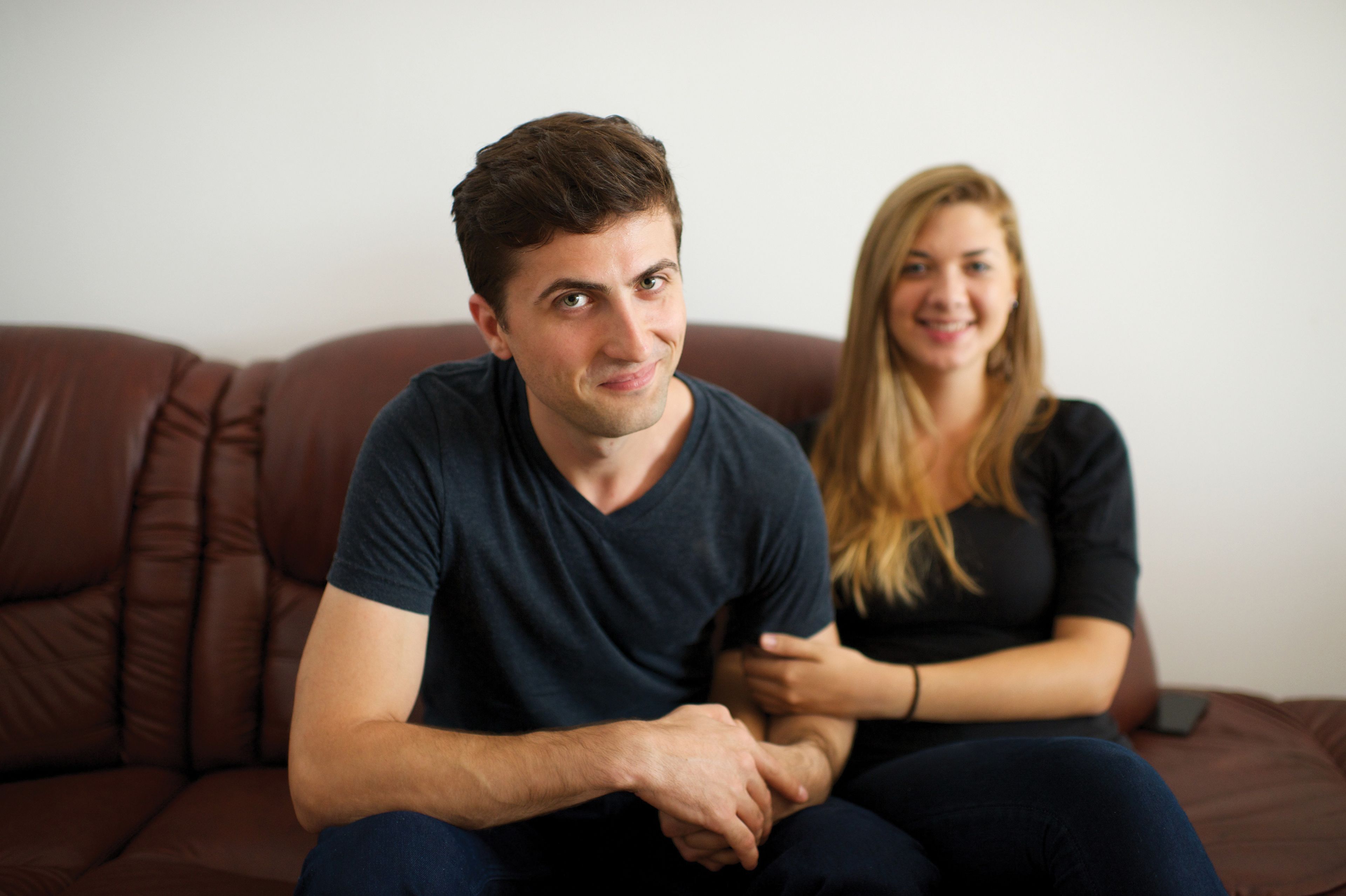 A young couple from Romania sitting on a brown couch beside each other.