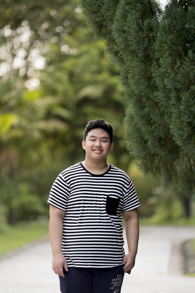 A young man stands in a park in Malaysia. He smiles to the camera.