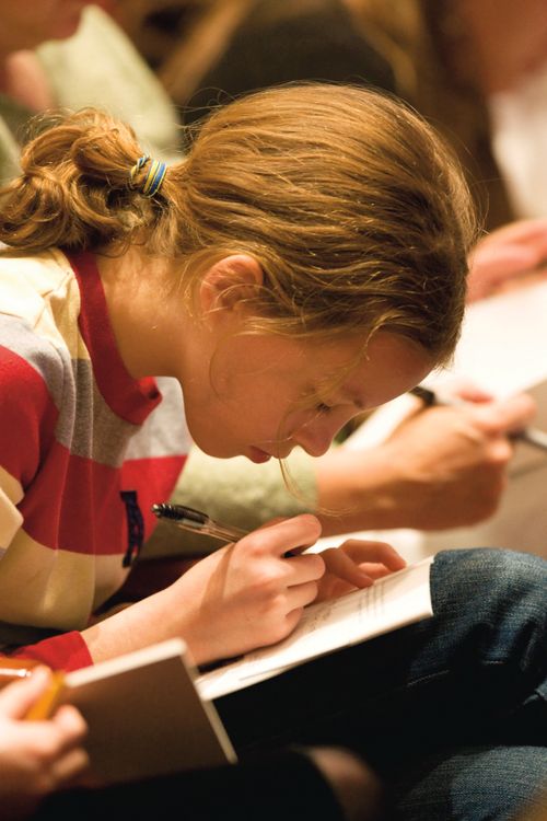 A girl with a light brown ponytail bends her head down as she writes in a notebook that is open on her lap.