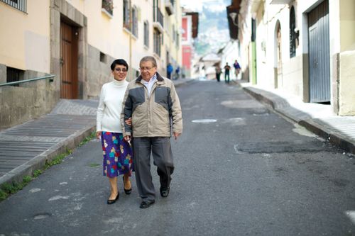 An elderly man in a jacket, pants, and dress shoes walks down a street arm-in-arm with his wife in a white sweater, floral skirt, and black shoes.