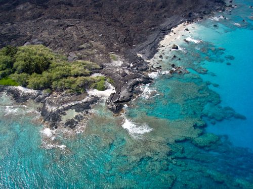 An aerial view of ocean water bordered by a rocky and green brush coastline in Hawaii.