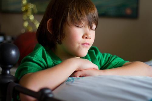 A young boy rests his folded arms on his bed, closes his eyes, and prays.
