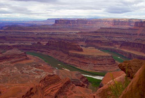 The dark green Colorado River runs through red rock canyons in southern Utah, with clouds covering the sky.