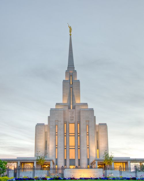 The front of the Oquirrh Mountain Utah Temple, with the lights on in the early evening and a row of colorful flowers on the grounds.