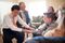 A blind young man shakes hands with family members during a ministering visit. His companion sits next to him with a happy family sitting near by.