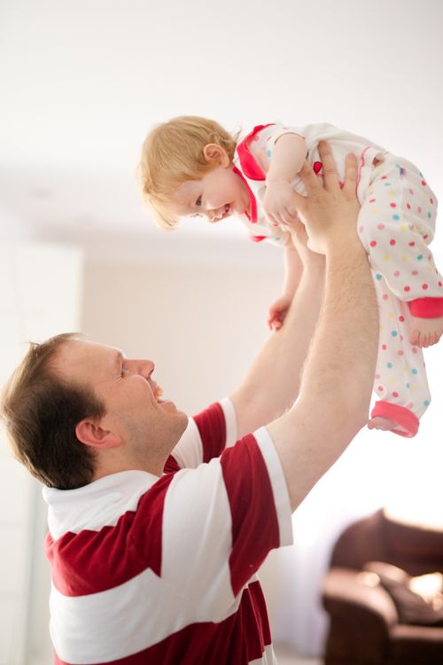 A man in a red and white striped shirt holds his baby girl high above his head and looks up at her smiling face.