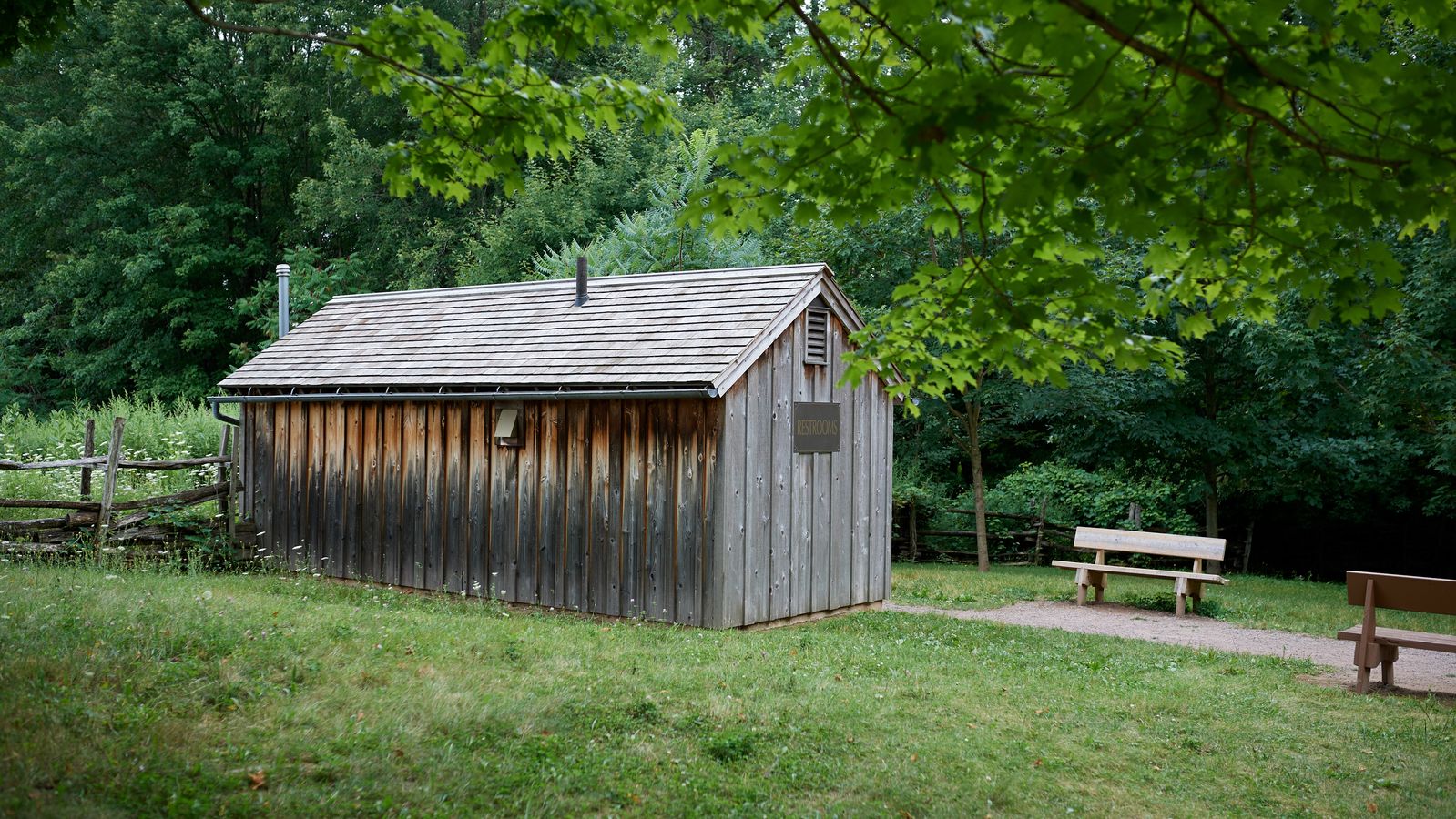 An exterior view of Joseph Smith's family exterior buildings in Palmyra, New York. 
There is farmland and a few smaller buildings on the land. They appear to be a barn and some kind of shed, likely another small barn or storage for a wagon.