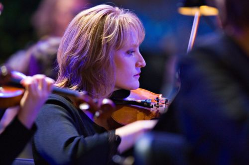 A woman with blonde hair and a black blouse plays a violin with other violinists in a concert.