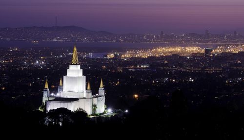 A view from afar of the Oakland California Temple, with the lights on in the evening and the glow of the city beyond.