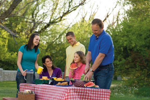 A father cuts a watermelon at a picnic table outside while the mother and their three children sit around the table.