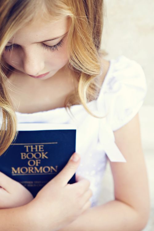 A young blonde girl wearing a white dress and holding a blue paperback copy of the Book of Mormon.