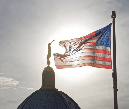 A photograph of the Tucson Arizona Temple dome and Angel Moroni statue with an American flag backlit by the sun.