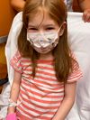 A little girl wearing a mask. Looks to be sitting on a hospital bed.