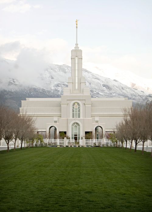 The front of the Mount Timpanogos Utah Temple, with a lawn and rows of trees leading to the entrance and with the mountain in the background.