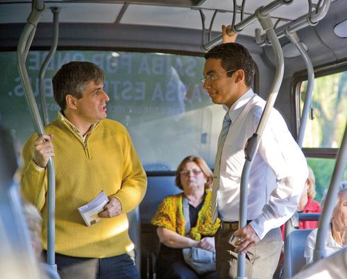 An elder missionary talking to a male passenger on a bus.