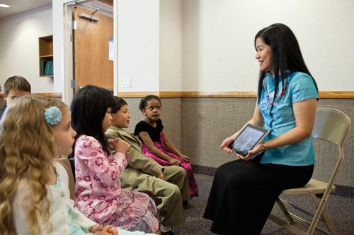 A Primary teacher with long black hair sits in a fold-out chair in front of five small children and shows them a picture of Christ on her iPad.