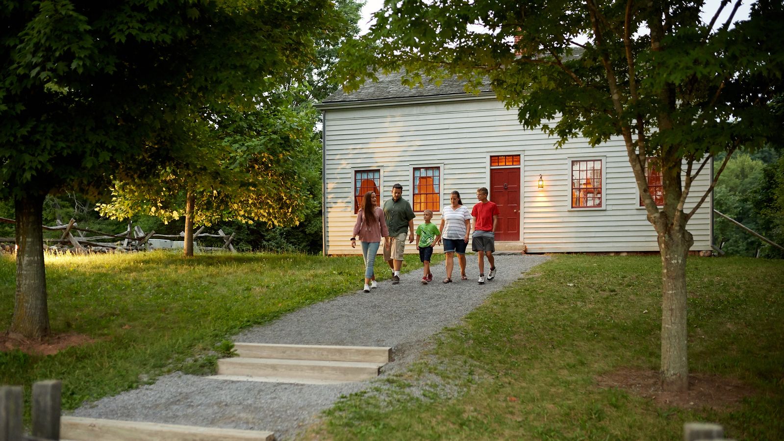 An exterior view of Joseph Smith's family home in Palmyra, New York. 
A family walks by the white building family farm. They are on paths around the farmland.