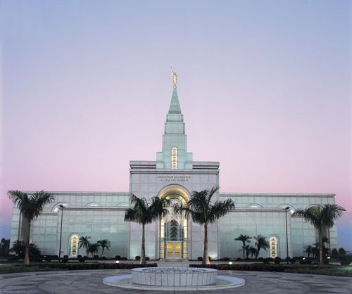 The Campinas Brazil Temple lit up in the early evening, with palm trees near the entrance.