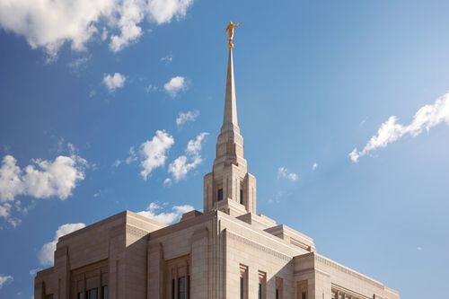 The top half of the Ogden Utah Temple and spire against a bright blue sky with small white clouds.
