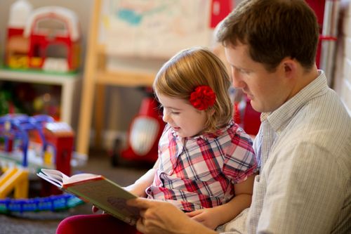 A father sits with his young daughter in a playroom and reads her a book.