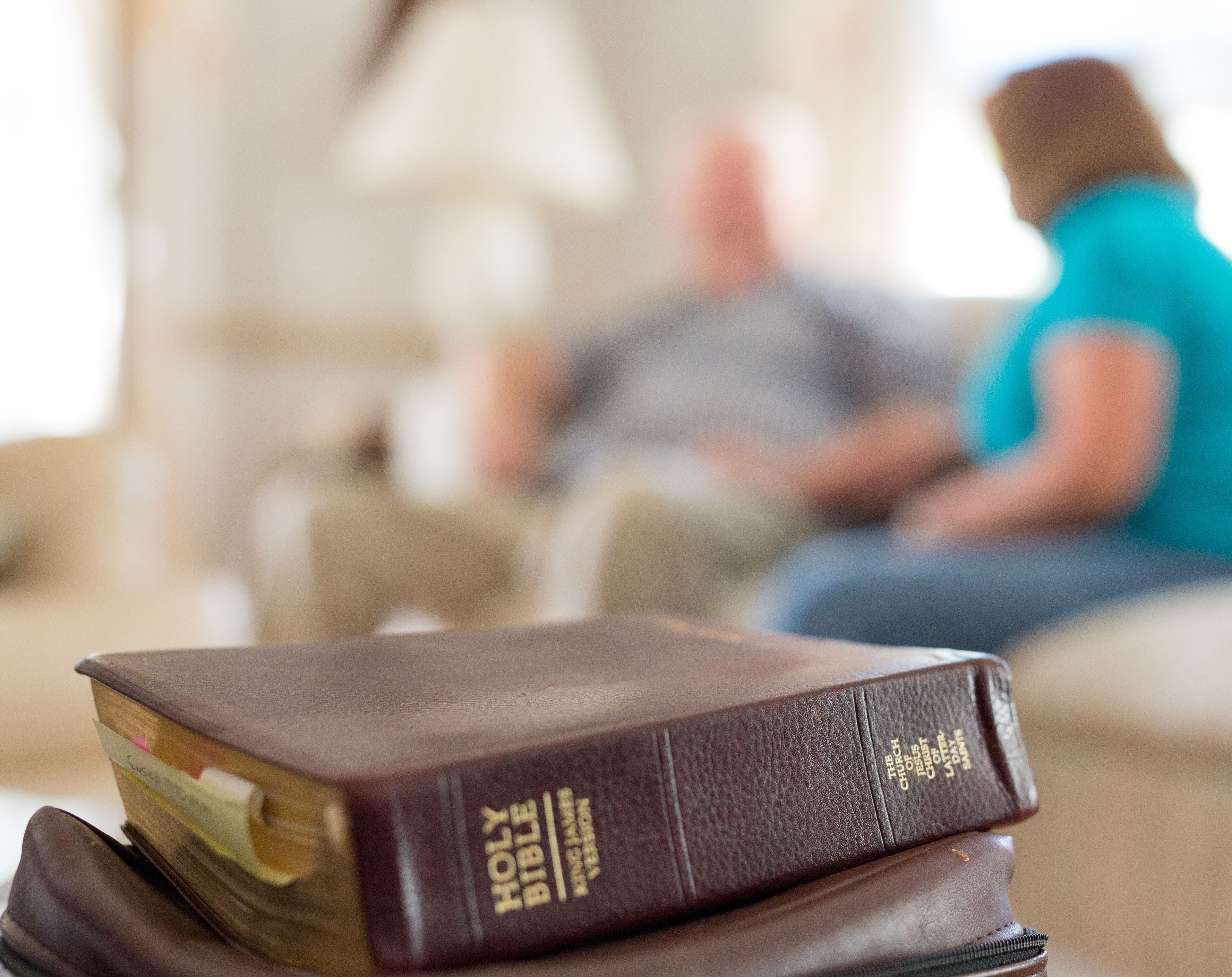 A Bible on top of a scripture case on a table, with a man and woman on a couch in the background.